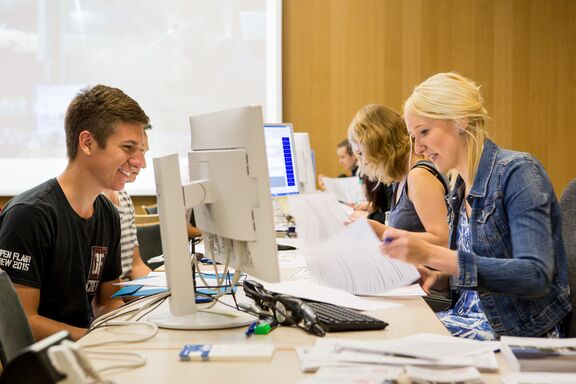 Mehrere Personen an einem Tisch. Im Vordergrund rechts eine weibliche Person, welche Unterlagen prüft, vor ihr auf dem Tisch ein Computer. Auf der linken Seite sitzt eine männliche Person vor dem Tisch und wartet. Im Hintergrund eine ähnliche Situation.