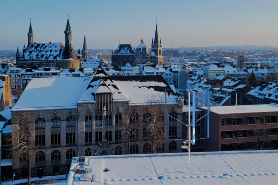 Foto von Aachen mit der Altstadt im Hintergrund. Im Vordergrund ist ein Modell des Vertikalwindrades auf ein Dach fotomontiert.