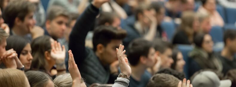 Ein Foto eines voll besetzten Hörsaals, mehrere Personen melden sich. Das Foto ist im Hintergrund unscharf.