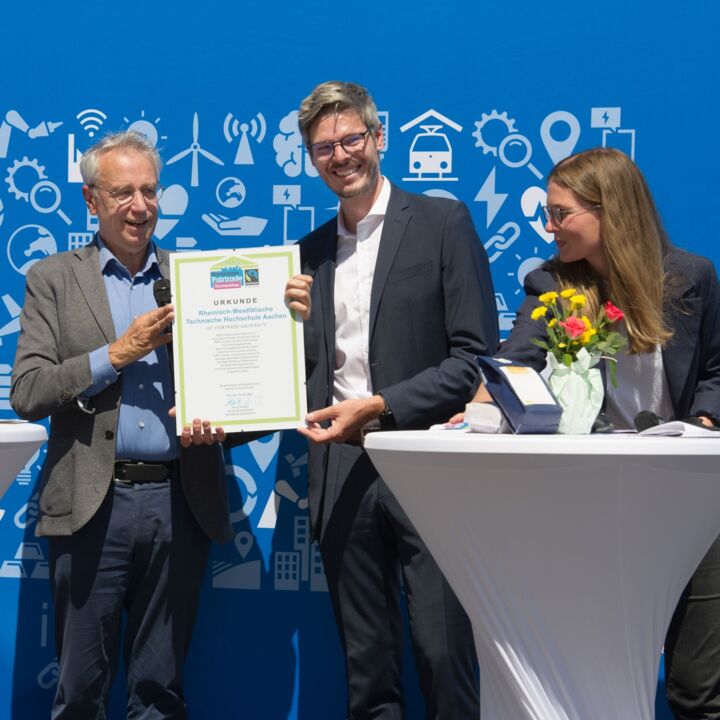 Three people stand in front of a blue photo wall with RWTH illustrations. Two people are jointly holding a certificate recognizing the university as a Fairtrade University. A third person looks at the certificate while leaning on a high table.