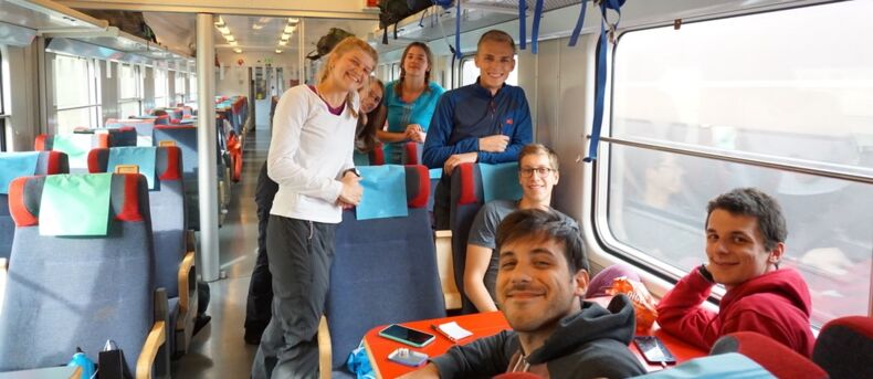 Das Bild zeigt sieben Personen in einem Zug. Drei Personen sitzen an einem Tisch. Vier Personen stehen. Alle lächeln in die Kamera.