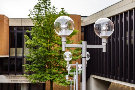 Das Foto zeigt die Straßenlaternen, die ausgehend vom Platanenplatz gegenüber des Hauptgebäudes der RWTH Aachen zum Karmanauditorium hinunter führen. In der Mitte des Fotos ist eine T-förmige Straßenlaterne abgebildet mit zwei kugelförmigen Lampen rechts und links oben. Die Leuchtmittel in den Lampen sind LEDs. Im Hintergrund sieht man drei weitere Straßenlaternen sowie das Karman-Auditorium und einen Baum.