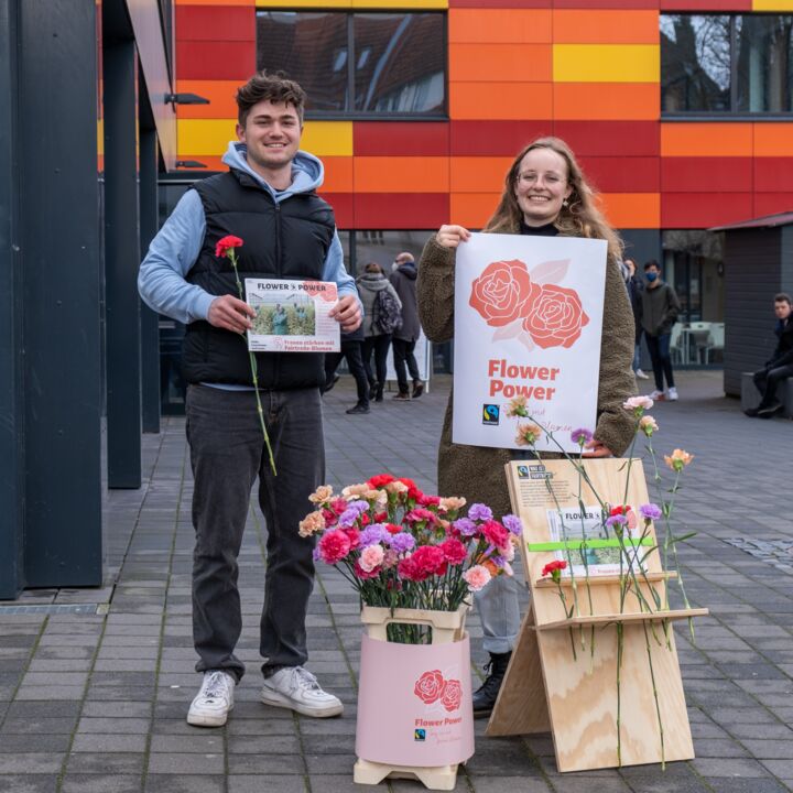 Two students stand in front of Mensa Academica and smile into the camera. The person on the left is holding a flower and an A4-sized information sheet about the Flower Power campaign. The person on the right is holding a larger poster with two roses and the words Flower Power. In the foreground are two wooden holders with many colorful flowers in them. Among them are red, purple and pink-beige blossoms.