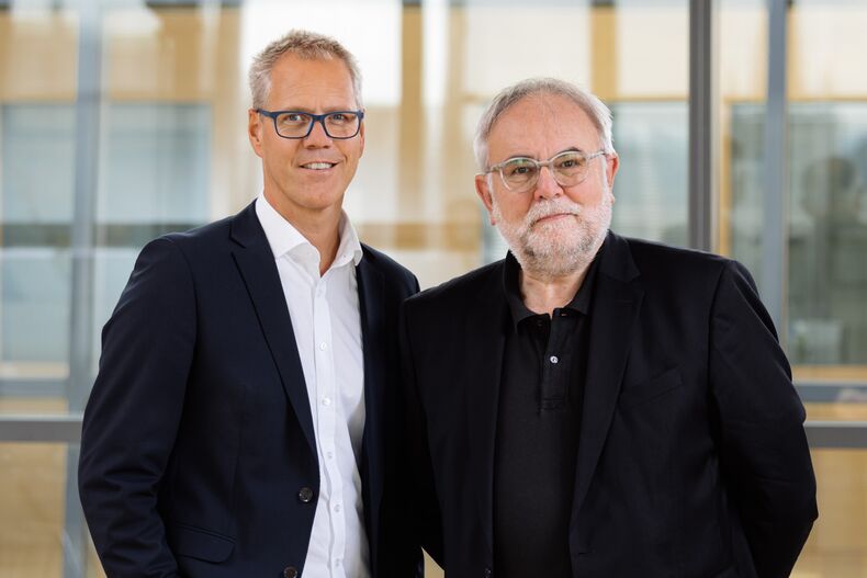 Foto des ständigen Kanzlervertreters Thomas Trännapp und des Kanzlers Manfred Nettekoven vor einer Glaswand. Thomas Trännapp mit schwarzem Sakko, weißem Hemd und Jeans. Manfred Nettekoven mit dunkelblauem Sakko, schwarzem Poloshirt und Jeans. Beide lächeln in die Kamera.