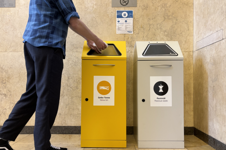 Two waste bins are next to each other, a yellow one on the left and a gray one on the right. A person throws a plastic bag into the yellow bin.