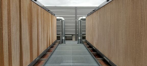Foto Adiabatisches Rückkühlwerk für den Hochleistungsrechner CLAIX.  Das Foto zeigt eine Konstruktion rechts- und linksseitig eines Gitterlaufweges. Am Ende führen zwei Rohre jeweils rechts und links in das Rückkühlwerk. Das Kühlwerk an sich ist von außen mit einer geriffelten Struktur in hellbraun sichtbar.
