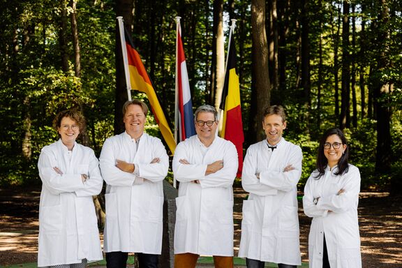 Zwei weibliche und drei männliche Personen in weißen Laborkitteln stehen vor den Flaggen von Deutschland, den Niederlanden und Belgien am Dreiländereck. Sie haben die Arme verschränkt und lächeln in die Kamera.