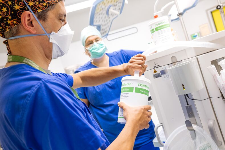 Eine weibliche und eine männliche Person stehen in einem Operationssaal. Sie tragen blaue OP-Kleidung, medizinische Schutzmasken und Hauben. Im Vordergrund schließt die männliche Person einen zylindrischen Behälter mit Narkosemedikamenten mit einem Schlauch an eine Maschine an.