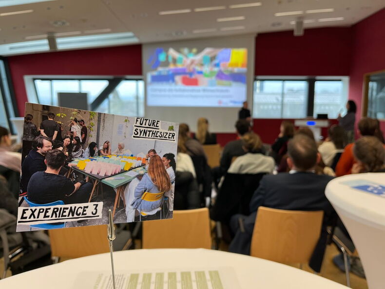 Im Vordergrund steht eine Postkarte auf einem Tisch, im Hintergrund sitzen Menschen auf Stühlen und hören einem Vortrag zu.