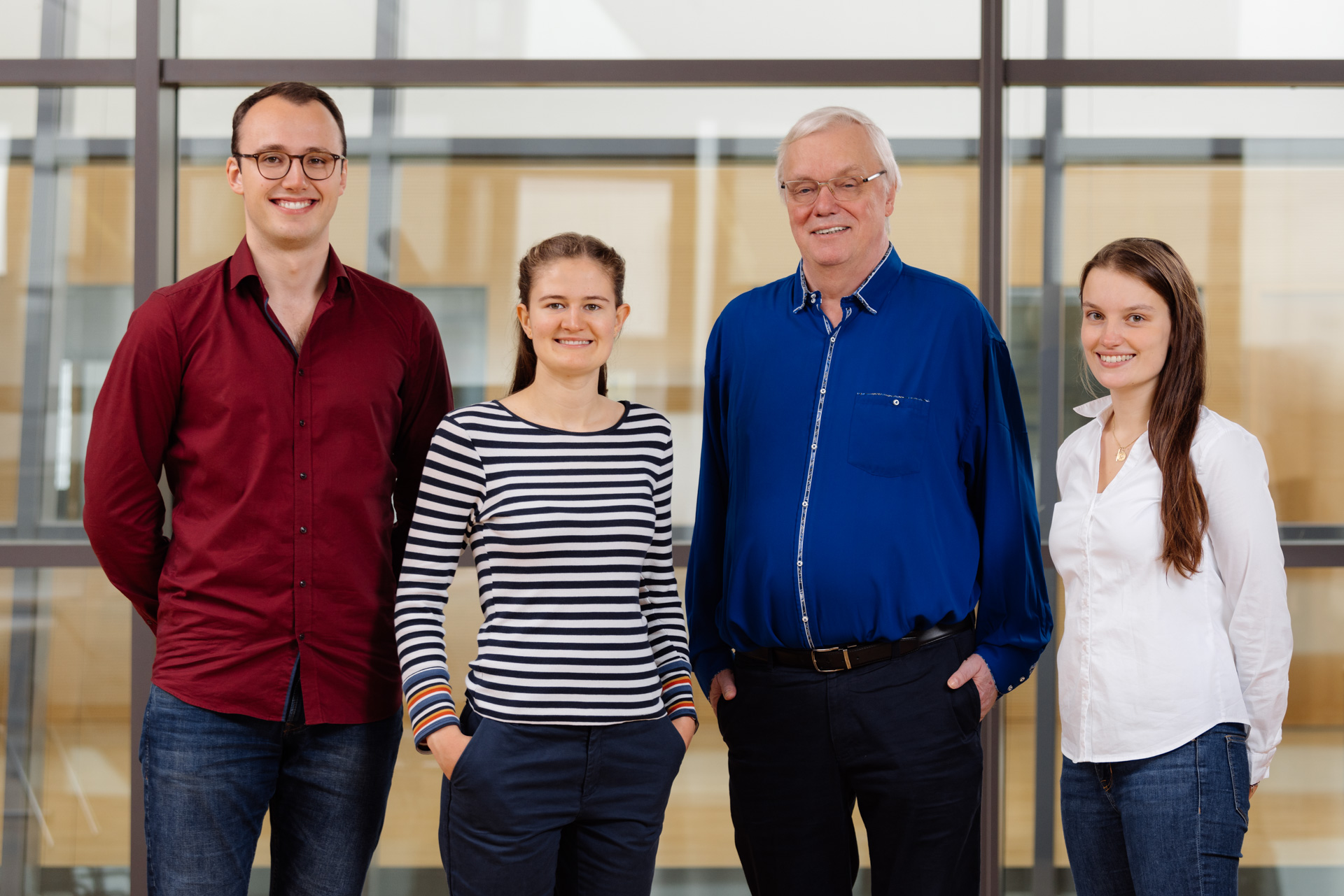 Ehemaliger AStA Referent für Lehre und Hochschulkommunikation Paul Heuermann, AStA Referentin für Nachhaltigkeit und studentisches Engagement Theresa Janning, Prorektor für Lehre Professor Aloys Krieg und ehemalige AStA-Vorsitzende Lena Schrader stehen vor einer Glaswand. Paul Heuermann trägt ein rotes Hemd und Jeans, Theresa Janning trägt ein schwarz-weiß gestreiftes Sweatshirt und Chino-Hose, Professor Krieg trägt ein königsblaues Hemd und Jeans, Lena Schrader trägt eine weiße Bluse und Jeans.  Sie lächeln in die Kamera.